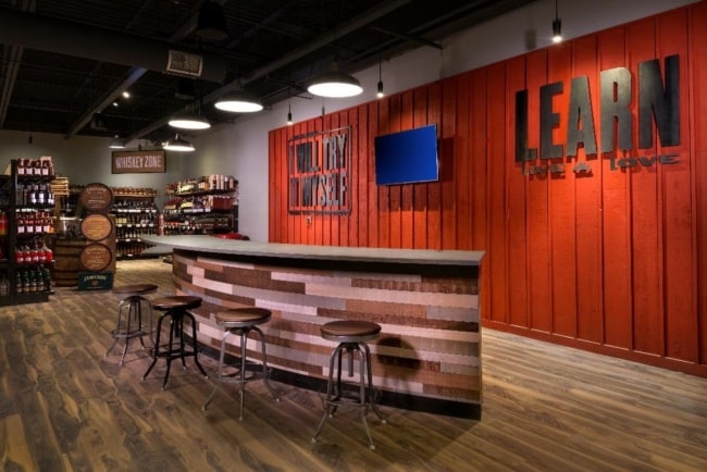 A section of the Chittenango liquor store is cleared of shelves, instead having a short bar with four stools on one side. A wooden paneled wall behind has a TV screen and "LEARN" in large letters.