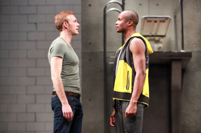 Two men and ex-lovers stand face-to-face on stage. Their hands are at their sides, but they are about to have a heated argument.