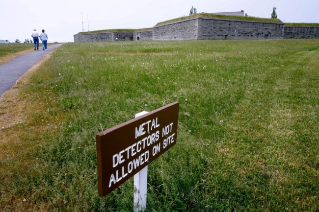 A fort sits at the top of a long, green lawn. A sign in the foreground reads "Metal detectors not allowed on site."