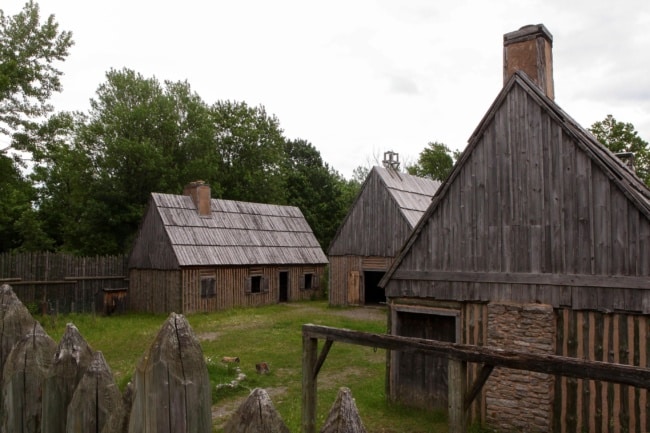 Three wooden buildings sit inside a fence off area made of sharpened wooden posts.