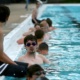 Teens line the side of a pool in the water, wearing goggles, training to be lifeguards.