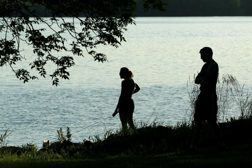 A man and a woman are silhouetted against the waters of Onondaga Lake as they stand at the shore. A small outline of a dog can just be seen past their feet in the tall grass.